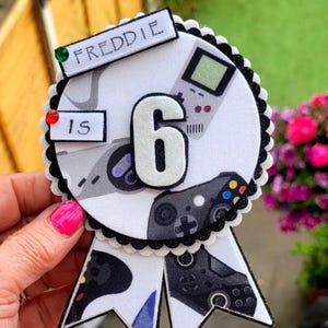 Peut inclure: Une rosette d'anniversaire sur le thème des jeux vidéo. La rosette blanche a un bord festonné noir et un grand chiffre 6 blanc. Elle présente des manettes de jeu et une console portable. Le nom "FREDDIE" est écrit sur une bannière.