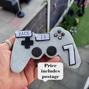 May include: A silver glitter-covered controller-shaped decoration with the text "JACK IS" and the number "7". The controller has black buttons and colourful accents. A speech bubble with the text "Price includes postage" is also visible.