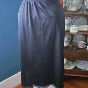 May include: A black slip skirt with an elastic waistband and a delicate lace trim at the hem. The skirt is displayed on a mannequin, showcasing its smooth, flowing fabric. The skirt is a classic piece of lingerie.
