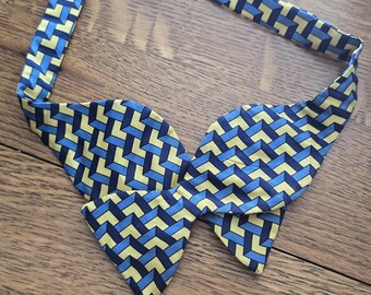 May include: A blue, yellow, and navy geometric patterned bow tie with a red adjustable neck strap. The bow tie is made of a silky material and is laid flat on a wooden surface. The bow tie is a classic accessory.