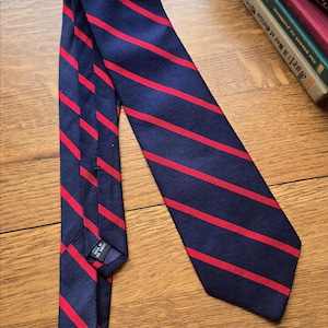 May include: Navy blue tie with diagonal red stripes. The tie is made of a textured fabric and is lying on a wooden surface. The tie is a classic accessory.