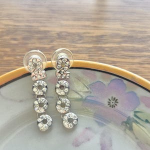 May include: Silver-toned dangle earrings with the number "2000" spelled out in clear, sparkling rhinestones. The earrings are displayed on a decorative plate with a floral design, showcasing the jewelry's elegant style.