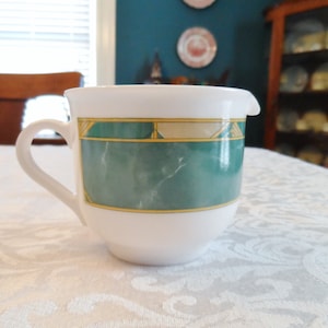 May include: White ceramic creamer with a green and gold geometric design. The creamer has a handle and a spout.