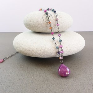 May include: A delicate necklace with a teardrop-shaped pendant made of pink sapphire. The necklace is made of silver chain and features small, colorful gemstones in shades of pink, blue, and orange.