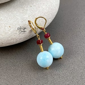 May include: Gold dangle earrings with a light blue round bead and a small red bead accent. The earrings have a lever back closure.