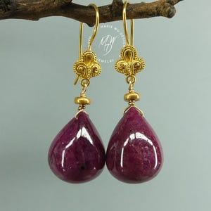 May include: A pair of gold dangle earrings featuring teardrop-shaped deep magenta gemstones suspended from ornate gold filigree tops. The earrings include a small gold bead spacer above each stone and a logo reading Marie Wu Design Jewelry.