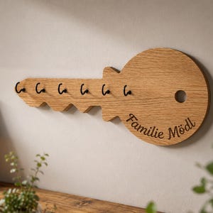 Könnte beinhalten: Ein wandmontierter Schlüsselhalter in Form eines großen Schlüssels aus hellbraunem Holz mit sichtbarer Maserung. Er verfügt über sechs schwarze Metallhaken an der Unterkante und den eingravierten Text "Familie Mödl" auf dem runden Kopf.