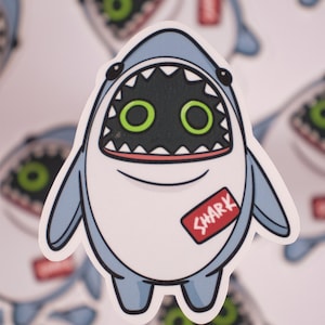 May include: A blue and white cartoon shark sticker with a big open mouth, green eyes, and a red label that says "SHARK".