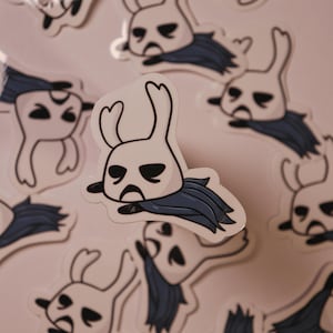 以下が含まれることがあります： 悲しい表情をした漫画のキャラクターが描かれた白と青のステッカーが複数あります。キャラクターは、角が2本とマントが付いた大きな頭をしています。ステッカーは薄いピンク色の背景に貼られています。