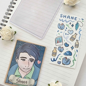 May include: A notebook with lined pages, a clear plastic cover, and a sticker sheet. The sticker sheet has the name "Shane" and illustrations of chickens, beer bottles, and other blue-themed graphics. A card with a cartoon portrait of a man is also present.