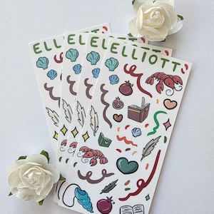 May include: A stack of stickers with the name "ELLIOTT" in green, featuring illustrations of seashells, lobsters, hearts, books, and other decorative elements. Two white paper roses are placed near the stickers.