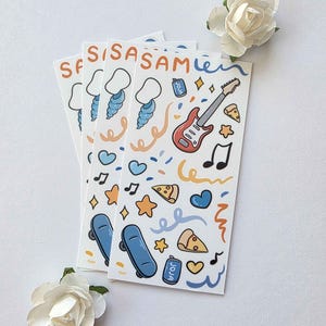 May include: Three white stickers with colorful illustrations of a guitar, pizza slices, skateboards, and musical notes. The stickers also feature the name "SASAM" and other decorative elements. Two white paper flowers are also in the image.