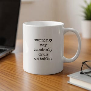 Puede incluir: Taza de cerámica blanca con asa negra. La taza tiene el texto "warning: may randomly drum on tables". La taza está sobre una superficie de madera.