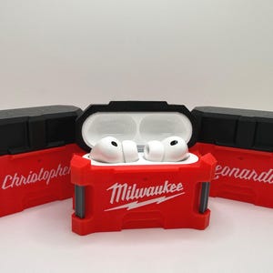 May include: Three red and black protective cases for wireless earbuds, designed to resemble tool boxes. The center case is open, revealing white earbuds inside. The cases feature the white text Milwaukee, Christopher, and Leonardo.