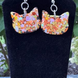 Può includere: Un paio di orecchini a forma di gatto con ganci argentati. Gli orecchini sono riempiti di glitter colorati, tra cui arancione, giallo e viola. Gli orecchini sono esposti su una superficie di velluto nero.