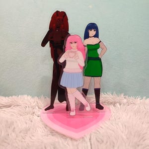May include: A decorative acrylic stand featuring three anime-style figures. The figures include a dark red character, a girl with pink hair, and a girl in a green dress. The stand is heart-shaped and pink.