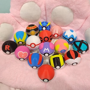 May include: A collection of colorful, spherical toys with various designs, including stripes, symbols, and geometric patterns. The toys are primarily red, blue, yellow, and pink, with white and black accents. The toys are arranged on a pink, plush surface.