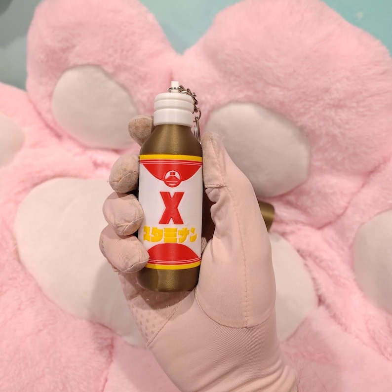 May include: A miniature bottle keychain with a gold body, white cap, and red and yellow label. The label features a large white "X" and Japanese text. The keychain is held against a pink background.