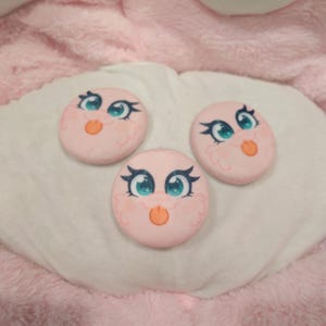 May include: Three pink, round, cartoon-style faces with blue eyes, orange beaks, and decorative eyelashes. The faces are arranged on a white, plush surface, with a pink background. The faces appear to be part of a larger plush toy or craft project.