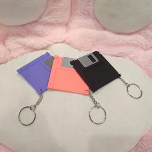 May include: Three colorful floppy disk keychains in purple, coral, and black. Each keychain has a silver-colored chain and a round key ring. The keychains are arranged on a white surface with a pink background.