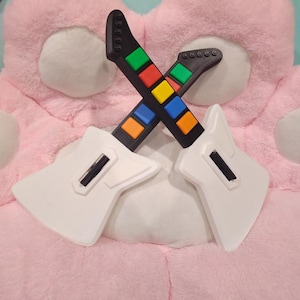 May include: Two toy guitars in a crossed formation. The guitars have white bodies and black necks with colorful buttons. The buttons are in the colors of orange, blue, yellow, green, and red. The guitars are resting on a pink surface.