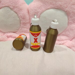 May include: Three gold-colored bottle keychains with white tops. One bottle has a label with a red and white design and the letter "X". The keychains are on a pink and white background.