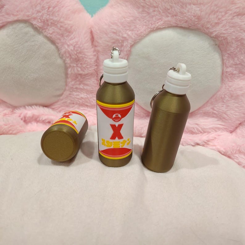 May include: Three gold-colored bottle keychains with white tops. One bottle has a label with a red and white design and the letter "X". The keychains are on a pink and white background.