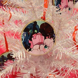 Puede incluir: Un adorno navideño redondo de madera con una ilustración de dibujos animados de personajes en colores azul, rosa y marrón. El adorno cuelga de un árbol de Navidad blanco con una cinta roja.