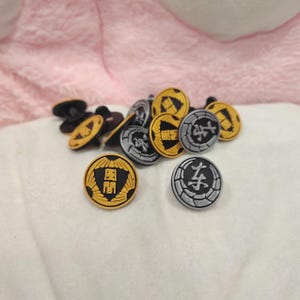 Op de afbeelding: Een verzameling ronde, decoratieve knopen met Japanse karakters. De knopen hebben zwarte, gouden en zilveren kleuren. De knopen liggen verspreid op een witte ondergrond.