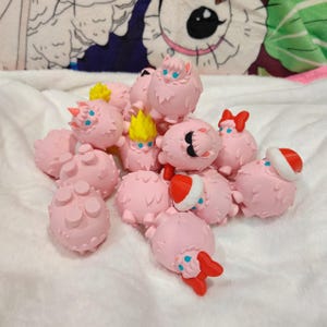 May include: A group of pink, spherical figurines with assorted accessories like red bows, Santa hats, and yellow hair. Some have blue eyes and small details. The figurines are arranged together on a white, textured surface.