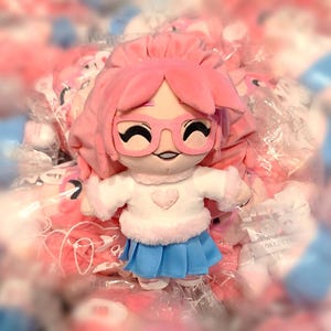May include: Pink plush toy with a white sweater, blue skirt, and pink hair. The toy is wearing glasses and has a heart on its sweater.