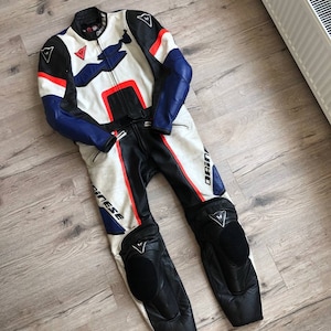 K&ouml;nnte beinhalten: Ein wei&szlig;er, blauer und roter Leder-Motorradrennanzug mit dem Dainese-Logo. Der Anzug hat Knie- und Ellbogensch&uuml;tzer.