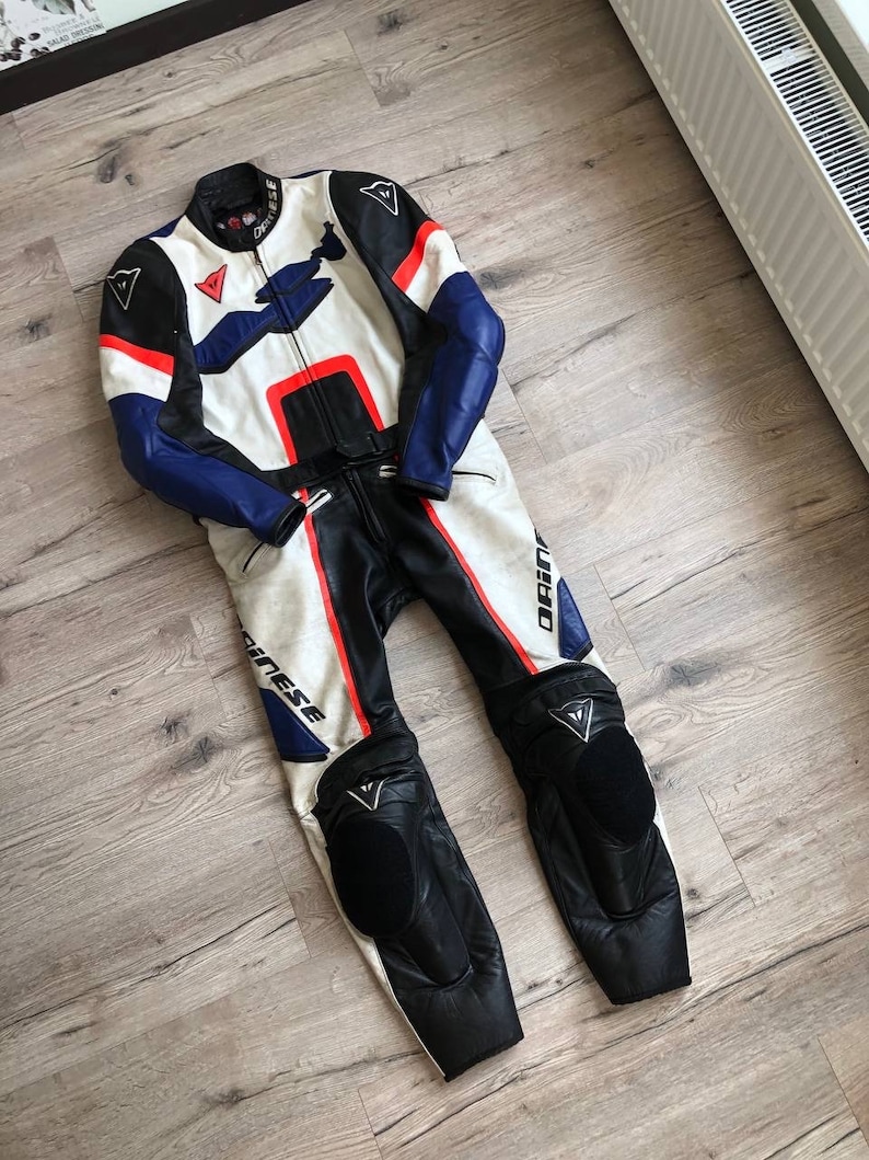 K&ouml;nnte beinhalten: Ein wei&szlig;er, blauer und roter Leder-Motorradrennanzug mit dem Dainese-Logo. Der Anzug hat Knie- und Ellbogensch&uuml;tzer.