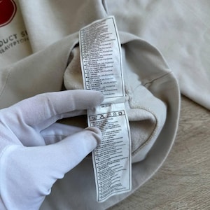 Op de afbeelding: Een wit sweatshirt met een rood Nike-logo en een label met wasinstructies. Het label bevat informatie over de stofsamenstelling, die een mix is van katoen en polyester.