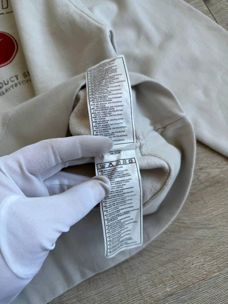 Op de afbeelding: Een wit sweatshirt met een rood Nike-logo en een label met wasinstructies. Het label bevat informatie over de stofsamenstelling, die een mix is van katoen en polyester.