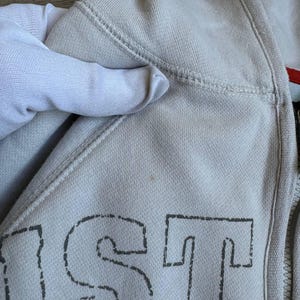 Op de afbeelding: Een lichtgrijs hooded sweatshirt met een zwart gestikt ontwerp dat "JUST" op de voorkant zegt.