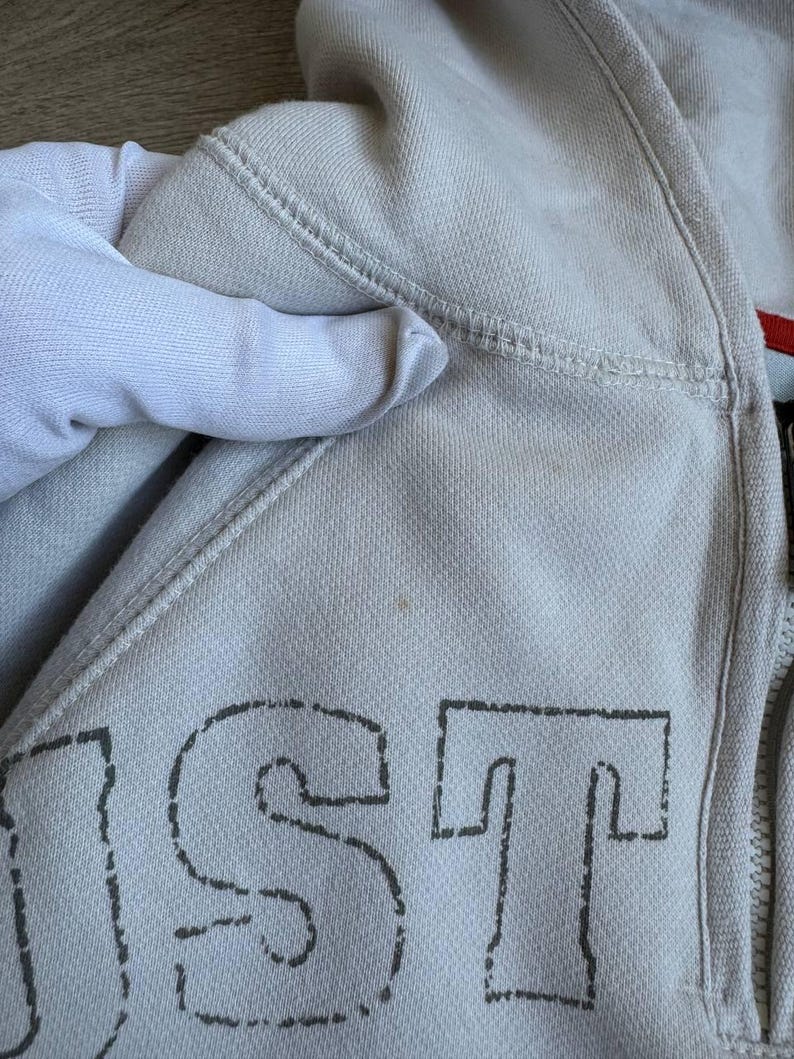 Op de afbeelding: Een lichtgrijs hooded sweatshirt met een zwart gestikt ontwerp dat "JUST" op de voorkant zegt.