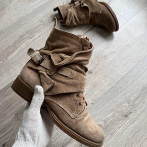 May include: A pair of tan suede ankle boots featuring a slouchy, folded upper design with a decorative side strap, a metal buckle, and brown laces. The boots have a low, flat heel and a rounded toe, showcasing a textured, matte finish throughout.