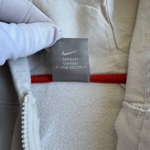 Op de afbeelding: Een wit hooded sweatshirt met een rode rits en een klein zwart label met de tekst "RN#56328 CA#05553  RFC#NM-920220KL4" aan de binnenkant van de capuchon.