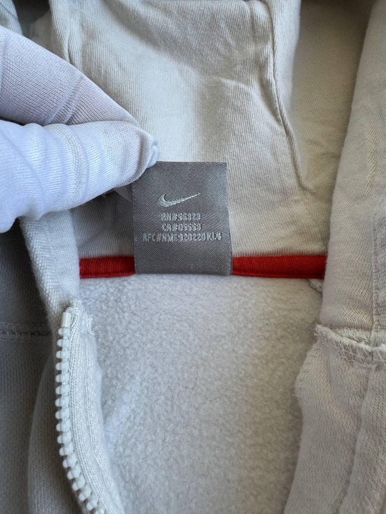 Op de afbeelding: Een wit hooded sweatshirt met een rode rits en een klein zwart label met de tekst "RN#56328 CA#05553  RFC#NM-920220KL4" aan de binnenkant van de capuchon.