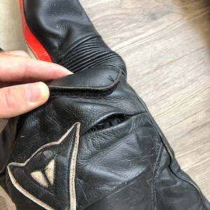 K&ouml;nnte beinhalten: Nahaufnahme eines schwarzen Ledermotorradanzugs mit roten und wei&szlig;en Akzenten. Der Anzug hat ein Dainese-Logo am Knie.