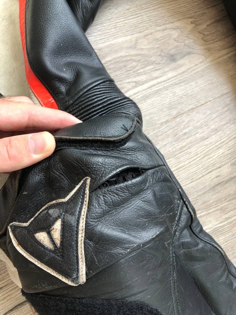 K&ouml;nnte beinhalten: Nahaufnahme eines schwarzen Ledermotorradanzugs mit roten und wei&szlig;en Akzenten. Der Anzug hat ein Dainese-Logo am Knie.