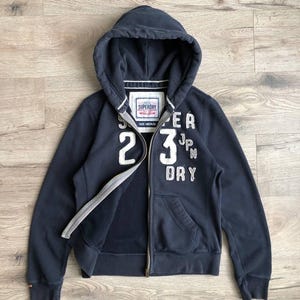 Peut inclure: Un sweat à capuche bleu marine zippé avec un logo Superdry blanc et le texte "Superdry 23 JPN DRY" sur le devant.