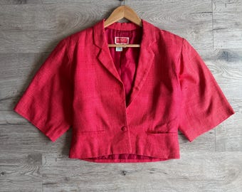 May include: A red cropped blazer with short sleeves, a notched collar, and a single-button closure. The blazer is hanging on a wooden hanger against a grey wood-grain background. The label inside the blazer reads "KENZO".