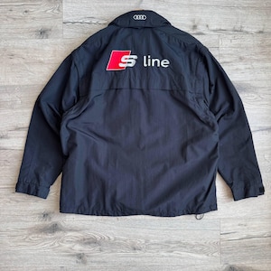 May include: Black jacket with the Audi logo and "S line" in white and red lettering on the back. The jacket has a collar and a drawstring at the bottom. The jacket is hanging on a metal hanger.