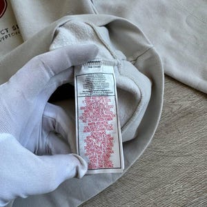 Op de afbeelding: Een wit sweatshirt met een rood logo en de tekst "CT SI" en "VERTOON". Het label op het sweatshirt heeft de tekst "KEEP AWAY FROM FIRE" en instructies in meerdere talen om het kledingstuk uit de buurt van warmtebronnen te houden.