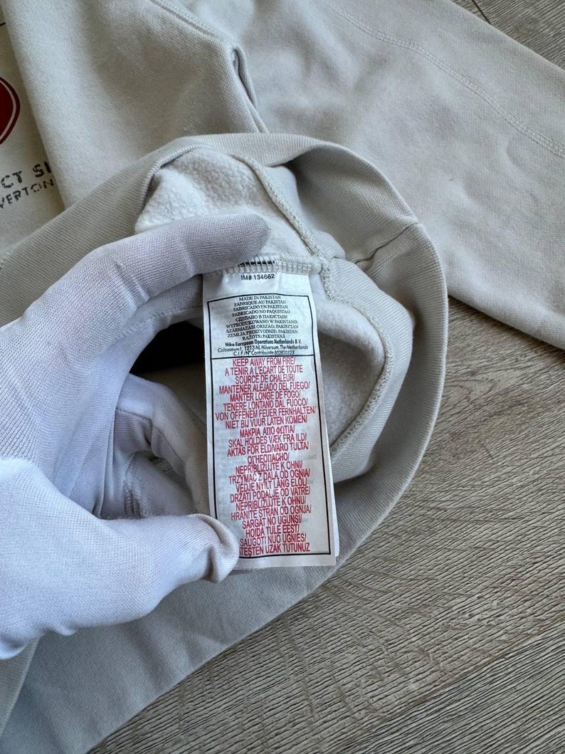 Op de afbeelding: Een wit sweatshirt met een rood logo en de tekst "CT SI" en "VERTOON". Het label op het sweatshirt heeft de tekst "KEEP AWAY FROM FIRE" en instructies in meerdere talen om het kledingstuk uit de buurt van warmtebronnen te houden.