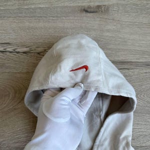 Op de afbeelding: Een lichtgrijs hooded sweatshirt met een rood Nike swoosh logo op de voorkant. De rits is zichtbaar aan de onderkant van het sweatshirt.