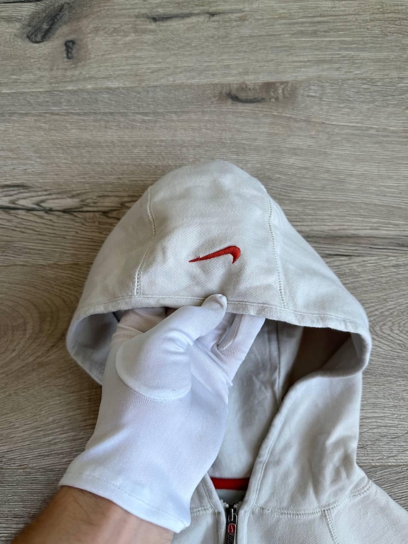 Op de afbeelding: Een lichtgrijs hooded sweatshirt met een rood Nike swoosh logo op de voorkant. De rits is zichtbaar aan de onderkant van het sweatshirt.