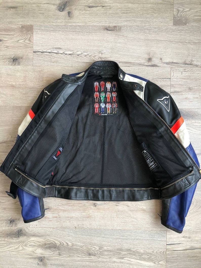 K&ouml;nnte beinhalten: Eine schwarze Leder-Motorradjacke mit blauen, wei&szlig;en und roten Akzenten. Die Jacke hat ein Mesh-Futter und ein Dainese-Logo auf der R&uuml;ckseite. Die Jacke ist ge&ouml;ffnet und das Futter ist sichtbar.